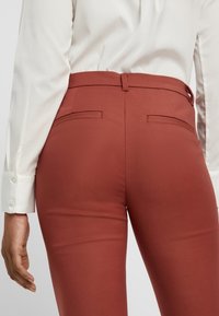 Rust-colored tailored pants with a smooth texture, featuring back pockets and a mid-rise waistband; paired with a white long-sleeve shirt.
