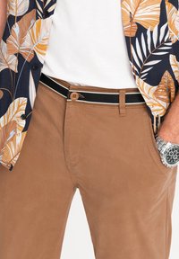 Chinos marroni con una texture liscia, caratterizzati da una cintura a righe bianche e nere. Abbinati a una camicia con una stampa tropicale multicolore.