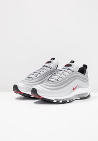 Stříbrné sportovní tenisky s vlnitým designem, černými a červenými akcenty, prodyšným síťovaným svrchním dílem a viditelným vzduchovým polstrováním v podrážce.