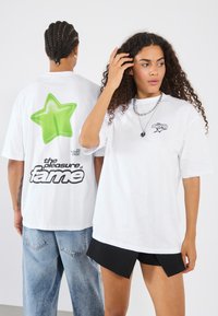 Camiseta de algodón blanca con gráfico de estrella verde en la parte posterior y texto "el placer de la fama", con un corte relajado y mangas cortas.