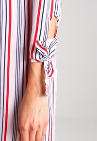 Raidallinen mekko, jossa on punaiset, valkoiset ja laivastonsiniset pystyraitat. Kangas näyttää kevyeltä, ja hihassa on solmu yksityiskohtia sekä aukkoja.