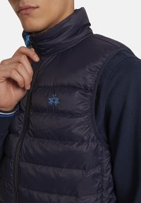 Navy sleeveless puffer vest with a high collar, featuring quilted sections, a zip closure, and a blue logo on the chest. Smooth, glossy texture.