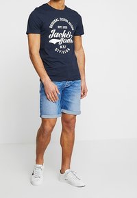 Hombre vestido con camiseta gráfica azul marino, pantalones cortos de mezclilla ligeros enrollados y zapatillas blancas, de pie frente a un fondo blanco.