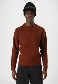Pull en tricot couleur rouille avec un col et des poignets côtelés, associé à un jean en denim noir. Un bonnet noir avec du texte blanc complète le look.