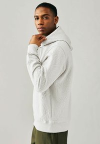 Sudadera con capucha de color gris claro, con un patrón floral texturizado, puños y dobladillo acanalados. El modelo se muestra de perfil, con un fondo neutro.