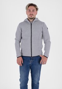 Felpa grigia con zip e design aderente, cerniera nera e cappuccio. Maniche lunghe con polsini neri, indossata sopra dei jeans blu. Tessuto semplice e strutturato.