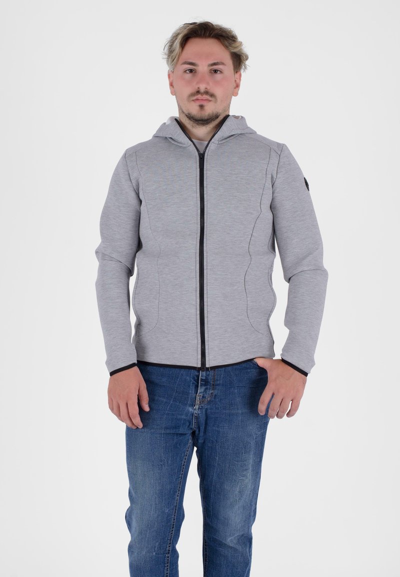 Felpa grigia con zip e design aderente, cerniera nera e cappuccio. Maniche lunghe con polsini neri, indossata sopra dei jeans blu. Tessuto semplice e strutturato.