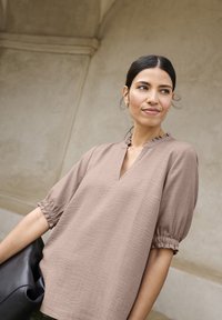 Blusa marrone a maniche corte con scollatura a V e colletto arricciato, realizzata in morbido tessuto testurizzato e con polsini elasticizzati. Il modello tiene in mano una borsa nera.
