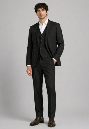 Jeune homme debout en costume trois pièces noir avec chemise blanche et chaussures marron sur fond gris uni.