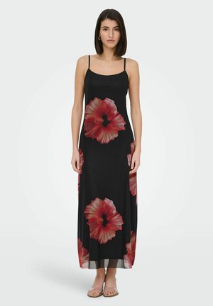 Kvinde iført en sort ærmeløs maxi-kjole med store røde blomsterprint, stående barfodet mod en ensfarvet lys baggrund.