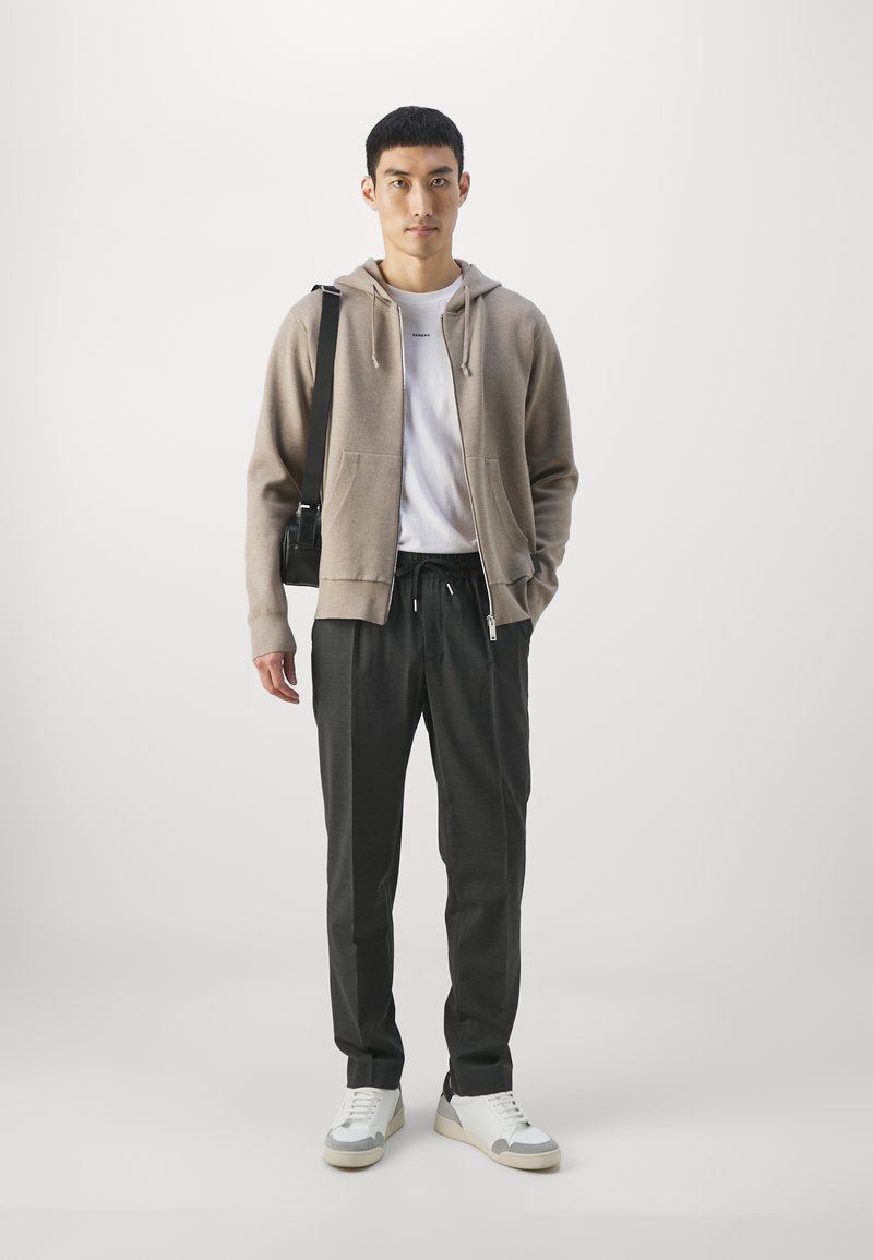 Beige Zip-Up-Hoodie über einem weißen T-Shirt, kombiniert mit dunklen Hosen und weißen Turnschuhen. Das Model hält eine schwarze Umhängetasche.