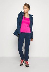 Γυναίκα με ναυτικό σακάκι και παντελόνι, φωτεινό ροζ μπλουζάκι και γκρι αθλητικά παπούτσια με ροζ κορδόνια, που στέκεται μπροστά σε απλό λευκό φόντο.