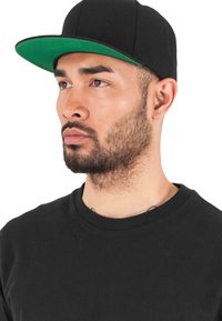 Must have mustaga, mille rohelise allpoole ja tasase visoriga, struktureeritud disain, kantud isiku poolt, kes kannab musta ümarat kraed T-särki valge tausta ees.