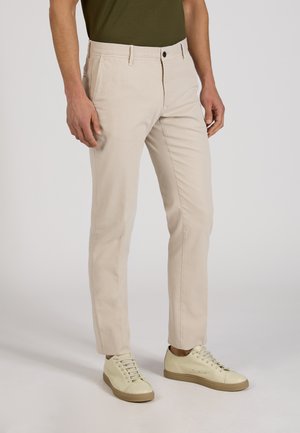 Mężczyzna w beżowych spodniach typu slim fit, kremowych sneakersach i ciemnozielonej koszuli, stojący na jasnym, jednolitym tle.