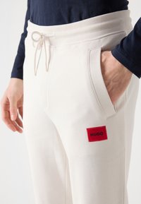 Pantalones de chándal crema con bolsillos laterales, cintura elástica y cordón. Presenta un parche rectangular rojo con "HUGO" en letras negras.
