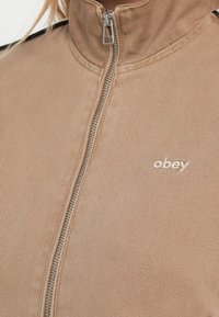 Jaqueta beige com fecho de correr prateado e pequeno logótipo "obey" bordado no peito.