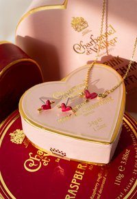 Ružová krabička v tvare srdca so zlatými detailmi obsahuje šperky: zlatý retiazkový náhrdelník a náušnice s červeným srdiečkom a motívom šípu.