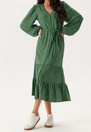 Femme portant une robe midi verte en œillets, manches longues bouffantes, boutonnée sur le devant, ceinture à la taille et baskets blanches, debout les mains sur les hanches.