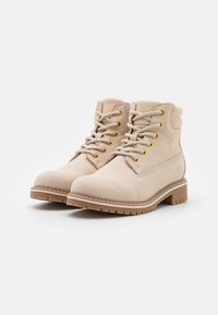 Bottines en daim beige avec un bout arrondi, des lacets beiges et des œillets dorés. Présente une semelle en caoutchouc texturée et un subtil détail de couture latérale.