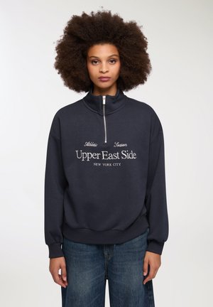 Felpa blu scuro con colletto alto, chiusura a mezza zip e testo bianco che recita "Upper East Side, New York City." Vestibilità morbida e rilassata.