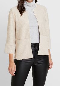 Beige texturovaná bunda s volným střihem, krátkým designem a 3/4 rukávy, spárovaná s šedým rolákem a přiléhavými černými kalhotami.