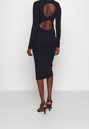 Vrouw draagt een nauwsluitende zwarte midi-jurk met lange mouwen en twee ovale uitsparingen op de rug, gecombineerd met zwarte sandaalhakken.