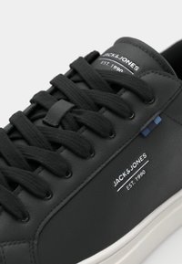 Baskets en faux cuir noires avec des lacets noirs, surface texturée, marquage blanc et un accent bleu près des œillets. Semelle extérieure blanche.
