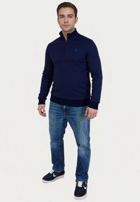 Marineblauwe zip-up trui met hoge kraag, textuurstof, gecombineerd met lichtblauwe jeans en marineblauwe sneakers met witte accenten.
