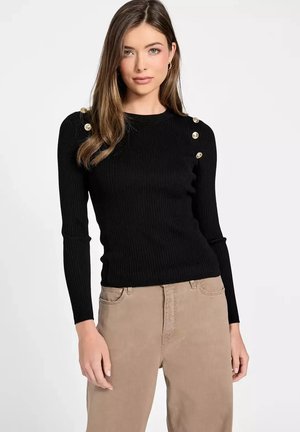 Joven mujer con un top negro de manga larga acanalado con botones dorados en los hombros y pantalones beige, de pie frente a un fondo liso.