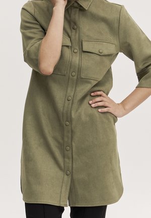 Mujer que lleva un vestido tipo camisa de manga larga en verde oliva con bolsillos de solapa en el pecho, una mano en la cadera y la otra cerca de su rostro.