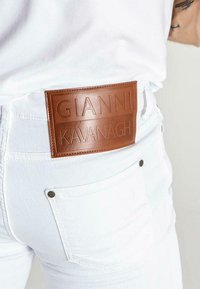 Valkoiset farkut, joissa on ruskea nahkainen bränditunnus "GIANNI KAVANAGH" takana. Varustettu metallinitoilla ja hienostuneella rakenteella.
