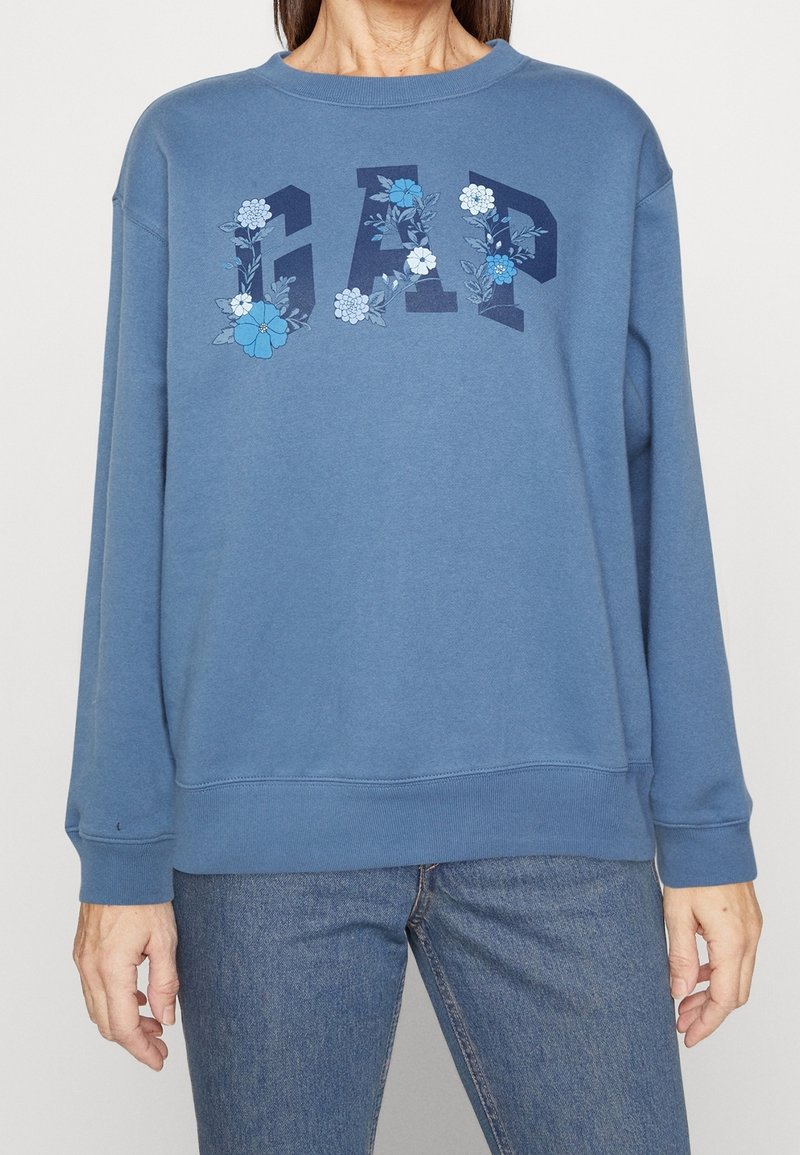Personne portant un sweat-shirt GAP bleu avec des motifs floraux à l'intérieur de lettres en gras, associé à un jean bleu moyen sur un fond uni.