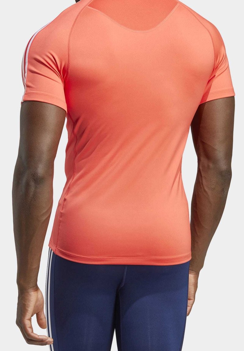 Camiseta deportiva de manga corta en un brillante color coral, hecha de una tela elástica, con costuras planas y un diseño ajustado. Acentuada con franjas blancas.