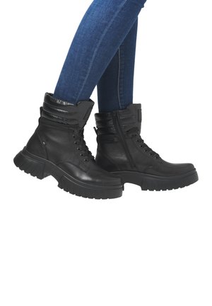 Botas de tobillo de cuero negro con suela robusta, cuello acolchado y detalle de cremallera. Combinadas con unos vaqueros ajustados azules, resaltando el diseño del calzado.