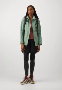 Nepremokavá mätovo zelená bunda, hnedý športový top, čierne šortky, čierne legíny a turistická obuv s červenými detailmi. Nastaviteľné ramenné popruhy.