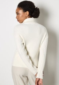 Maglione bianco a coste con collo alto e maniche a trecce, caratterizzato da un design aderente. Texture liscia con un aspetto complessivo morbido.