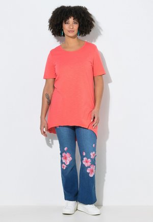 Kobieta o kręconych włosach stoi, ubrana w koralowy top z krótkim rękawem, niebieskie dżinsy z różowymi kwiatowymi wzorami, białe trampki i turkusowe kolczyki.