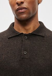 Καφέ πλεκτό πόλο πουκάμισο με υφή, με κολάρο και δύο μαύρα κουμπιά στον λαιμό. Χωρίς διακριτά μοτίβα ή αξεσουάρ.