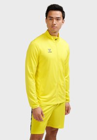 Mand iført en lys gul sportstrøje med lynlås og matchende shorts med en sort stribe på shorts, stående mod en ensfarvet baggrund.