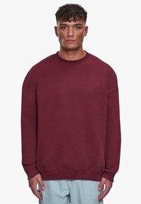 Burgunderfarbener Pullover aus weichem Material, mit Rundhalsausschnitt, überschnittenen Schultern und gerippten Bündchen. Lässige Passform mit einem Logo-Detail.