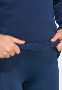 Felpa blu navy realizzata in tessuto testurizzato, con orlo a maniche corte. La persona tiene l'orlo con entrambe le mani, mostrando dei leggings blu navy attillati.