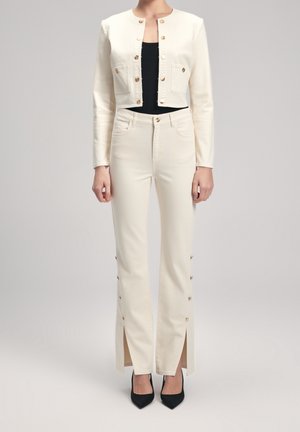 Veste crème cintrée avec des boutons dorés, associée à un pantalon crème taille haute avec des fentes latérales et des accents de boutons dorés. Haut noir et talons.