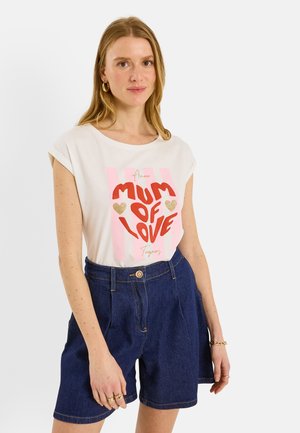 Femme aux cheveux clairs portant un t-shirt blanc à manches courtes avec le texte « Mum of Love » et un short en jean bleu taille haute, debout devant un fond blanc.