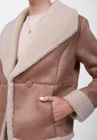 Brun kunstlærfrakk med en fluffy beige krage, som har en enkelt knappelukking og sidelommer, og som viser en myk tekstur og en strukturert design.