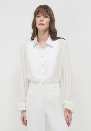 Woman wearing a sheer white blouse with a structured bib front and white high-waisted trousers against a plain white background.