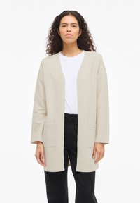 Μπεζ μακρύς ζακέτα με υφή, ανοιχτό μπροστά και δύο μπροστινές τσέπες, φορεμένο πάνω από λευκή μπλούζα και μαύρο παντελόνι.