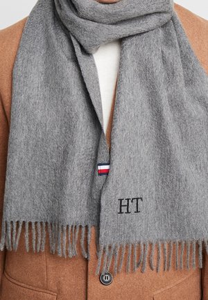Écharpe en laine grise avec bords à franges, initiales brodées "HT" et petite étiquette rouge, blanche et bleue près du bord. Portée sur un pull léger.