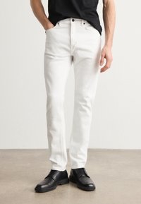 Witte denim broek met een slim fit, vijf zakken en metalen knopen. Draag met een zwart shirt en zwarte schoenen op een neutrale achtergrond.