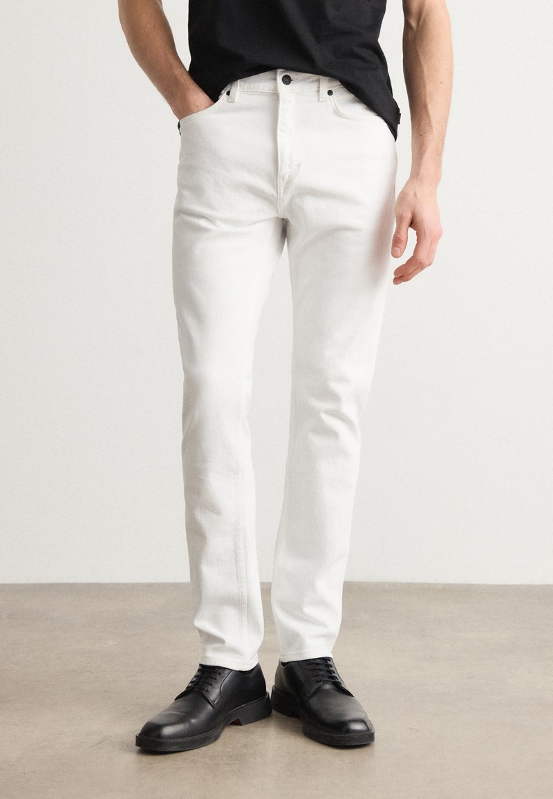 Witte denim broek met een slim fit, vijf zakken en metalen knopen. Draag met een zwart shirt en zwarte schoenen op een neutrale achtergrond.