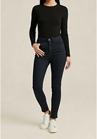 Woman wearing black long-sleeve top, high-waisted dark skinny jeans, and white sneakers, standing with one hand in pocket.